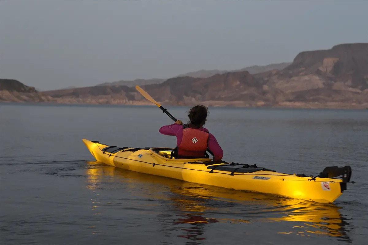 Person kayaking on a calm lake at sunset, with mountains in the background.