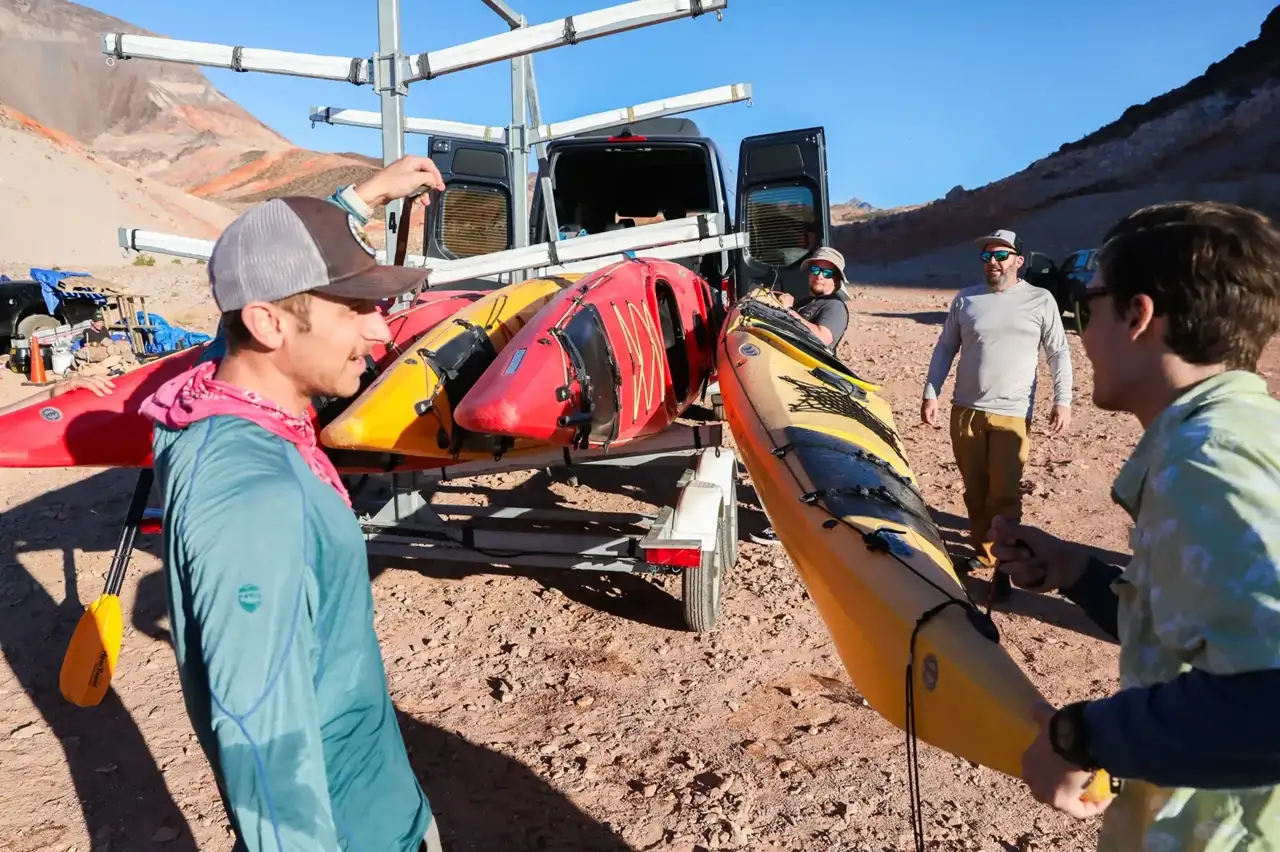 Group of people loading kayaks onto a trailer in a rocky, desert landscape.