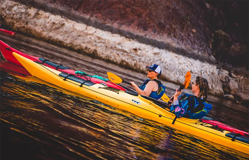 Two people kayaking on a river with rocky shorelines in the background.