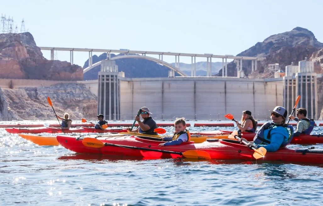 People kayaking on a lake with a dam and bridge in the background on a sunny day.