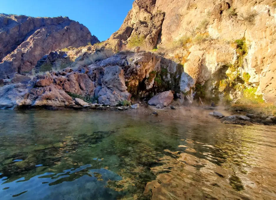 Rocky landscape with a clear water pond and steaming hot spring, under a blue sky.
