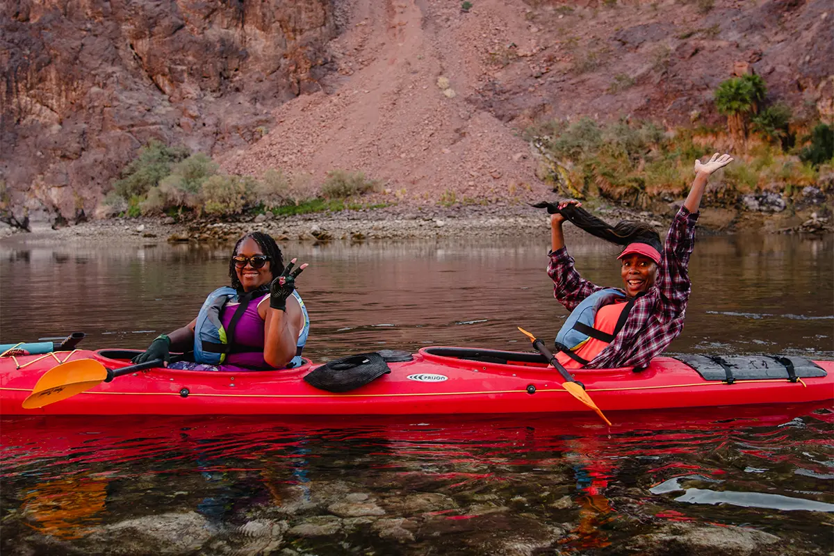 Two people in a red kayak smiling on a calm river with rocky cliffs behind them.