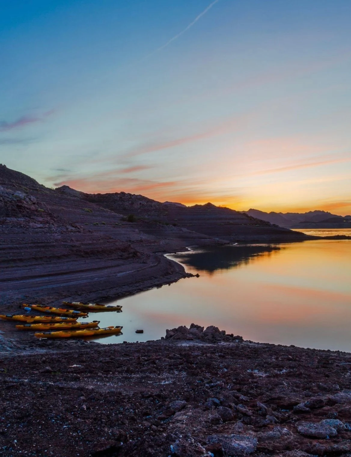 Sunset over a rocky shore with yellow kayaks and calm water reflecting colorful sky.