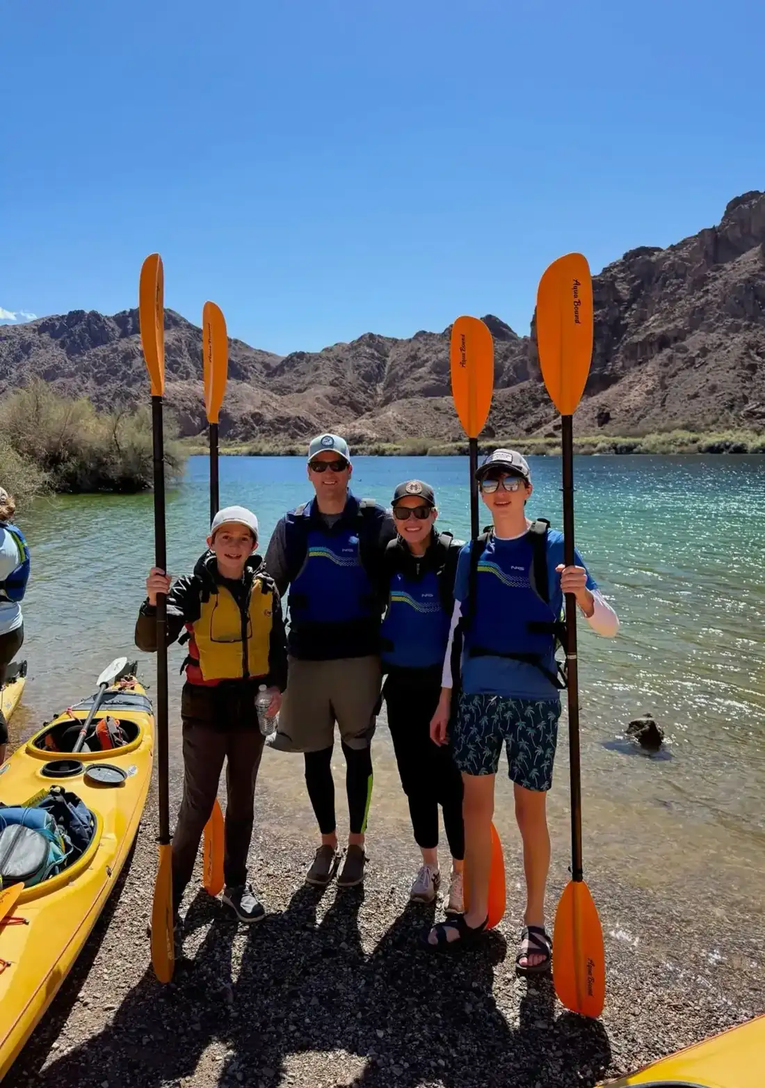Four people in life vests hold kayak paddles by a lake with mountains in the background.