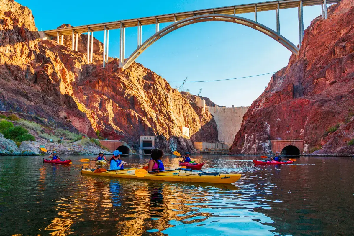 People kayaking on a river under a large bridge and between rocky cliffs.