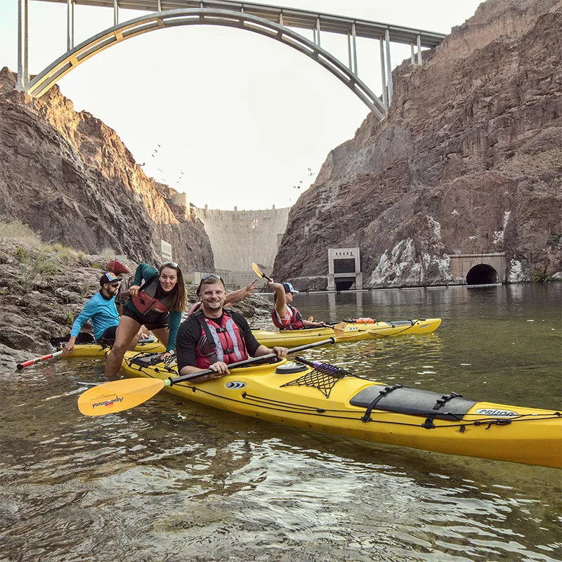 People kayaking on a river beneath a large arched bridge and rocky cliffs.