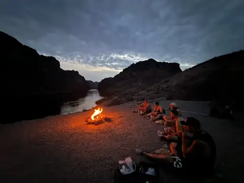 Group gathered around a campfire on the riverbank as dusk falls over the canyon