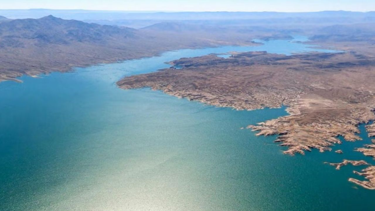 Aerial view of a winding, blue lake surrounded by arid, rocky terrain and distant hills.