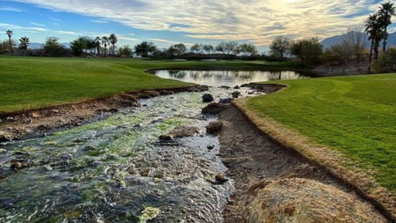 Scenic view of a golf course with a stream, pond, and palm trees under a cloudy sky.