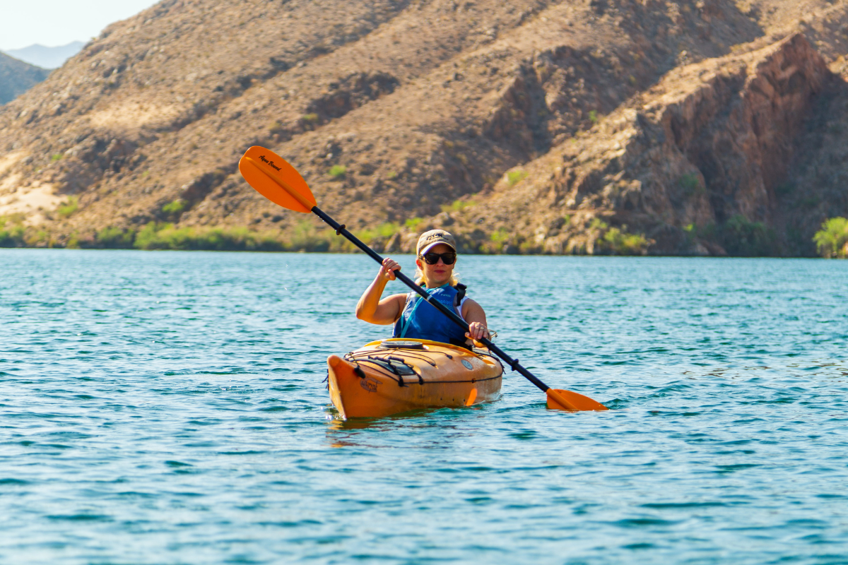 Person kayaking on a blue lake with rocky hills in the background.