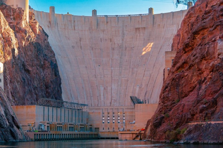 Large concrete dam with water reservoir, surrounded by rocky cliffs under a clear blue sky.