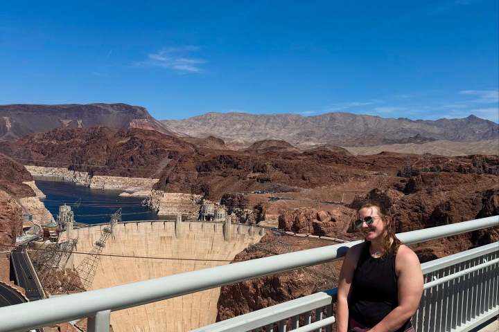 Person standing on a bridge railing with Hoover Dam and rocky landscape in background under clear blue sky.
