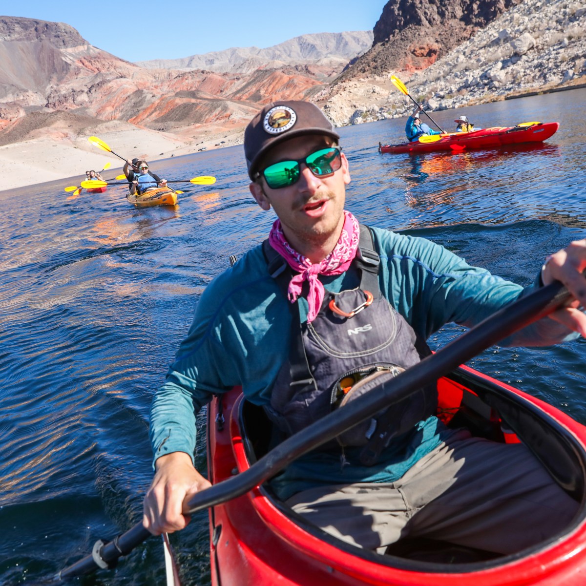 Man kayaking in foreground with two others in kayaks on a lake surrounded by rocky mountains.