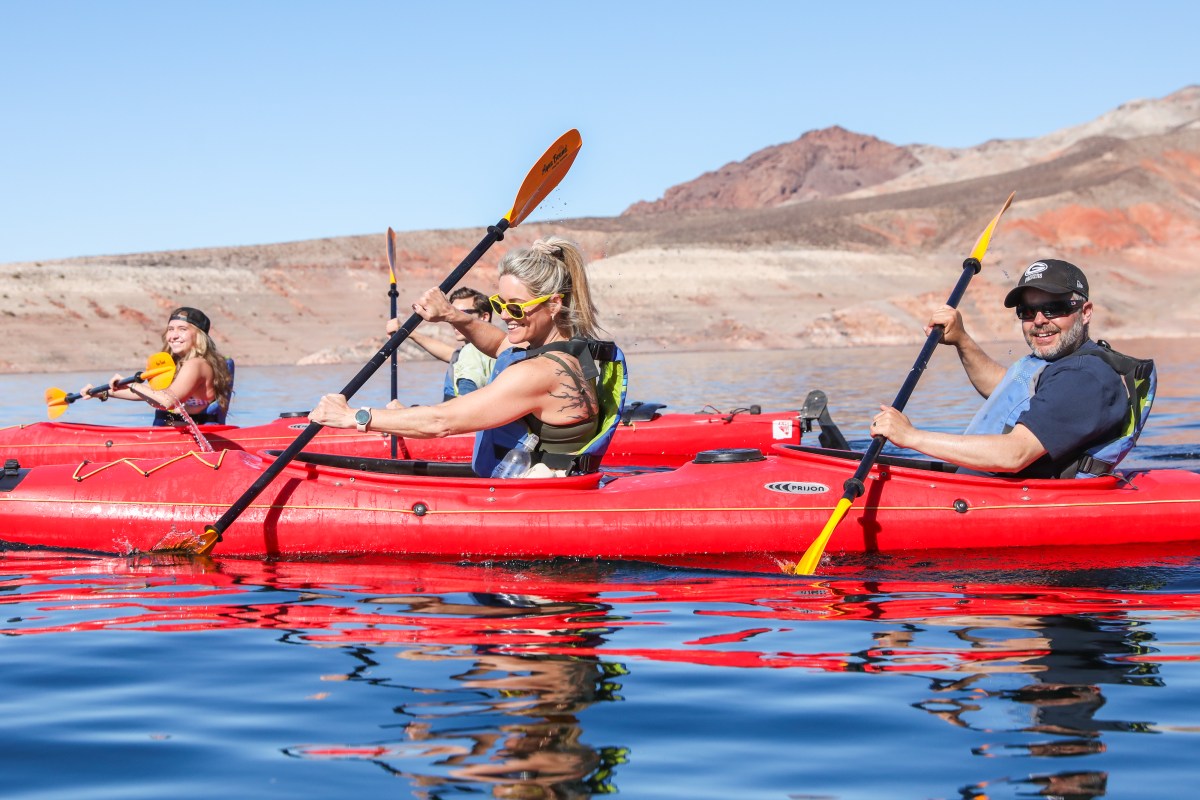 Four people kayaking in red kayaks on a lake with rocky hills in the background.