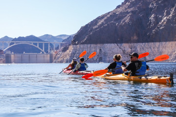 Group kayaking on a lake with mountains and bridge in the background.