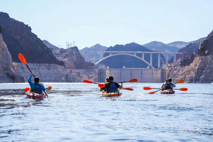 Three people kayaking on a lake near a large bridge and rocky mountains.