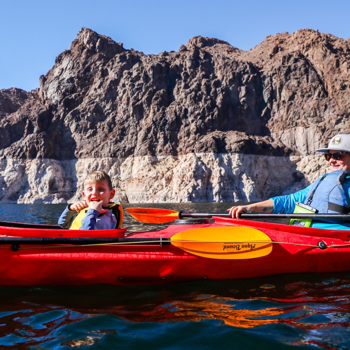 Two people kayaking on a lake with rocky cliffs in the background.