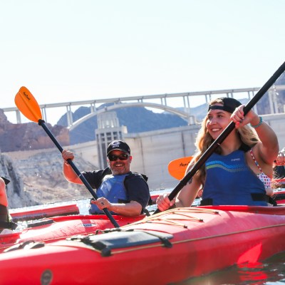Four people kayaking in red kayaks near a dam, wearing life jackets and smiling.