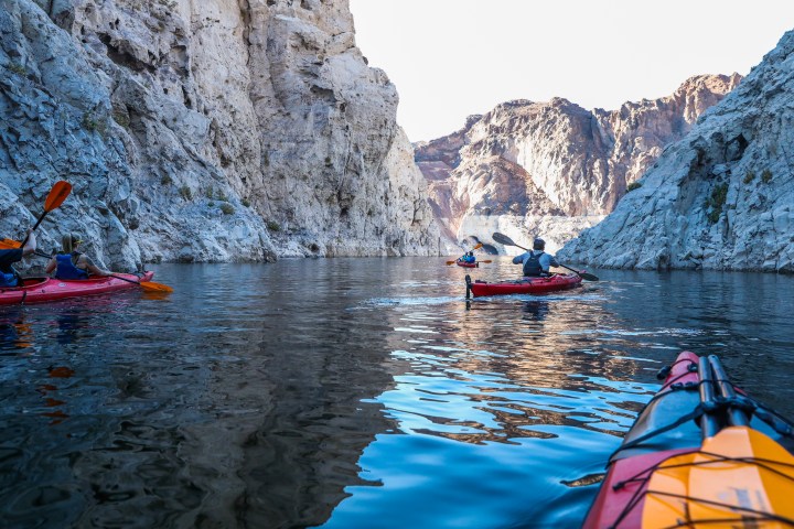 Kayakers paddle through a narrow, rocky canyon with calm water and sunlit cliffs.