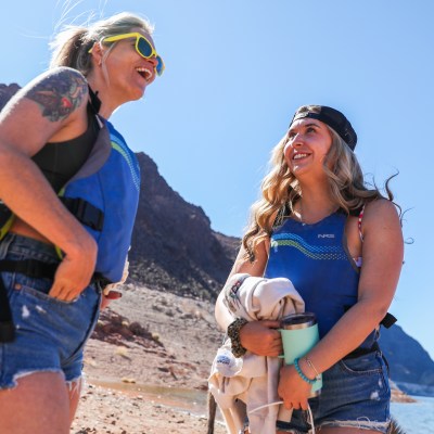 Two women laughing near a lake in life vests and shorts on a sunny day.