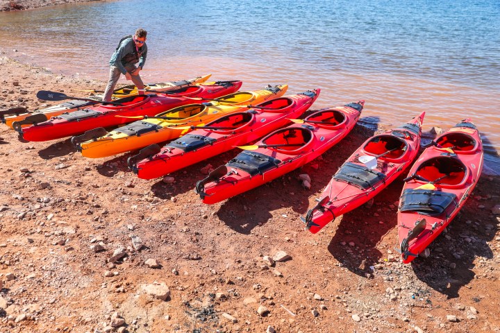 Person arranging colorful kayaks on a rocky lakeshore.
