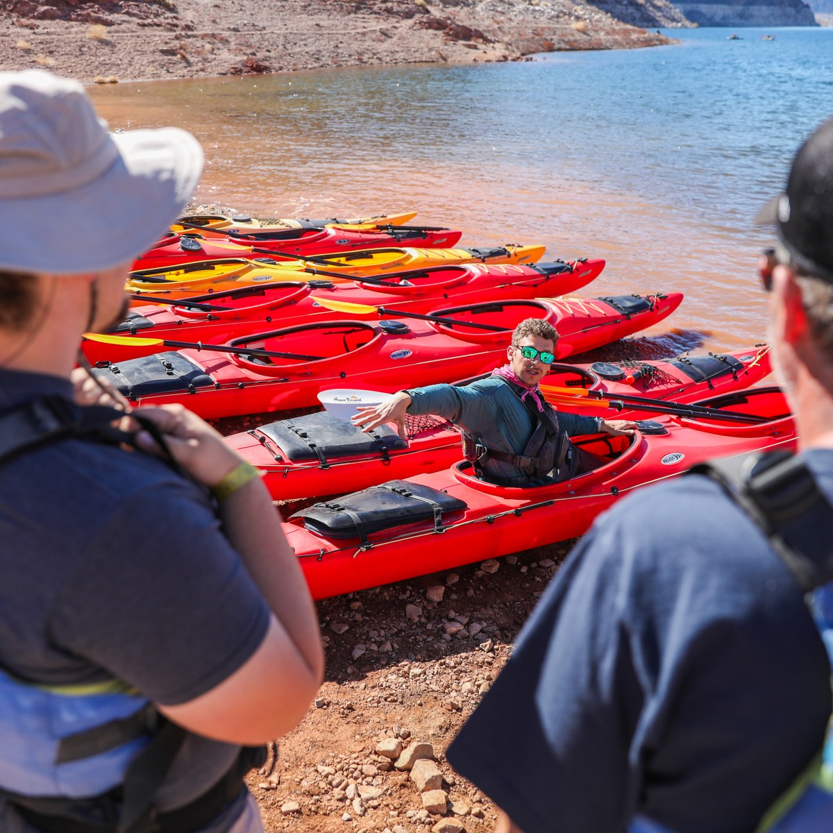 Group listening to a person in a kayak at the edge of a lake with red cliffs.