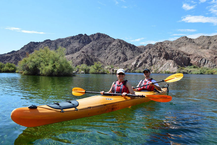 Two people kayaking on a calm lake with rocky hills in the background under a blue sky.