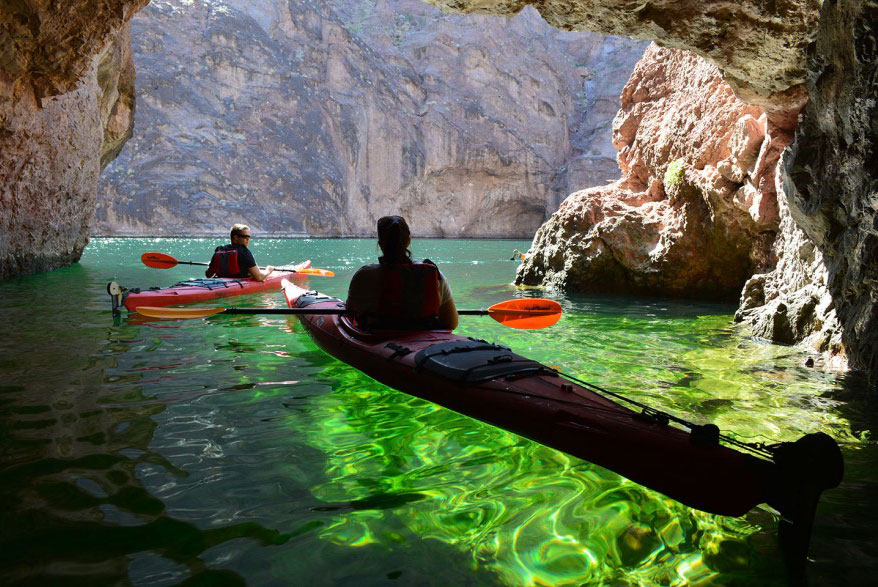 Two kayakers paddle through a rocky cave with sunlight illuminating green water.