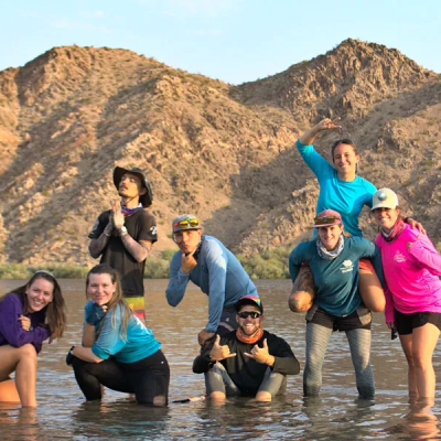 Group of people posing playfully in water with rocky hills in the background.