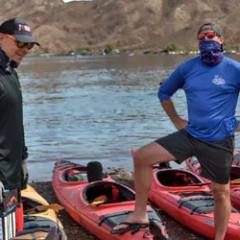 Two people wearing sun protection gear stand by red kayaks on a rocky shore.