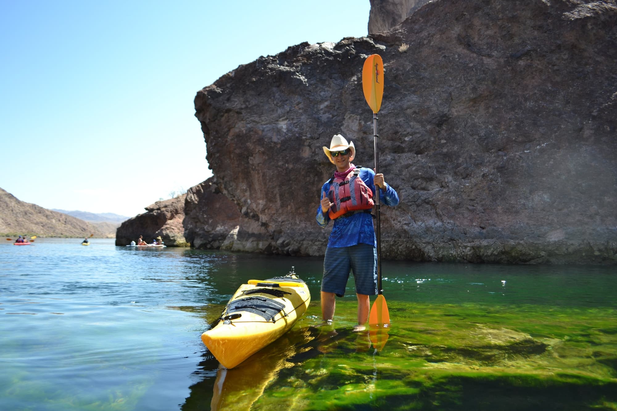 Person in cowboy hat stands beside a yellow kayak on clear water, holding a paddle near rocky cliffs.