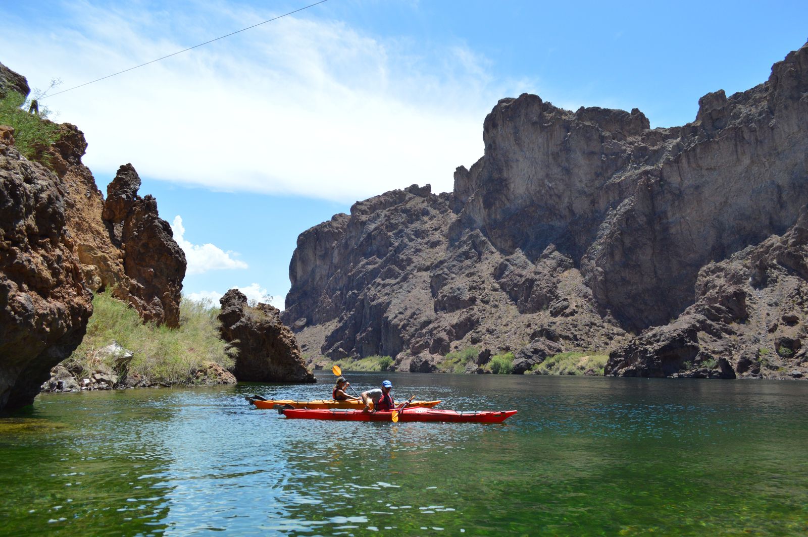Two people kayaking on a river surrounded by rocky cliffs under a clear blue sky.