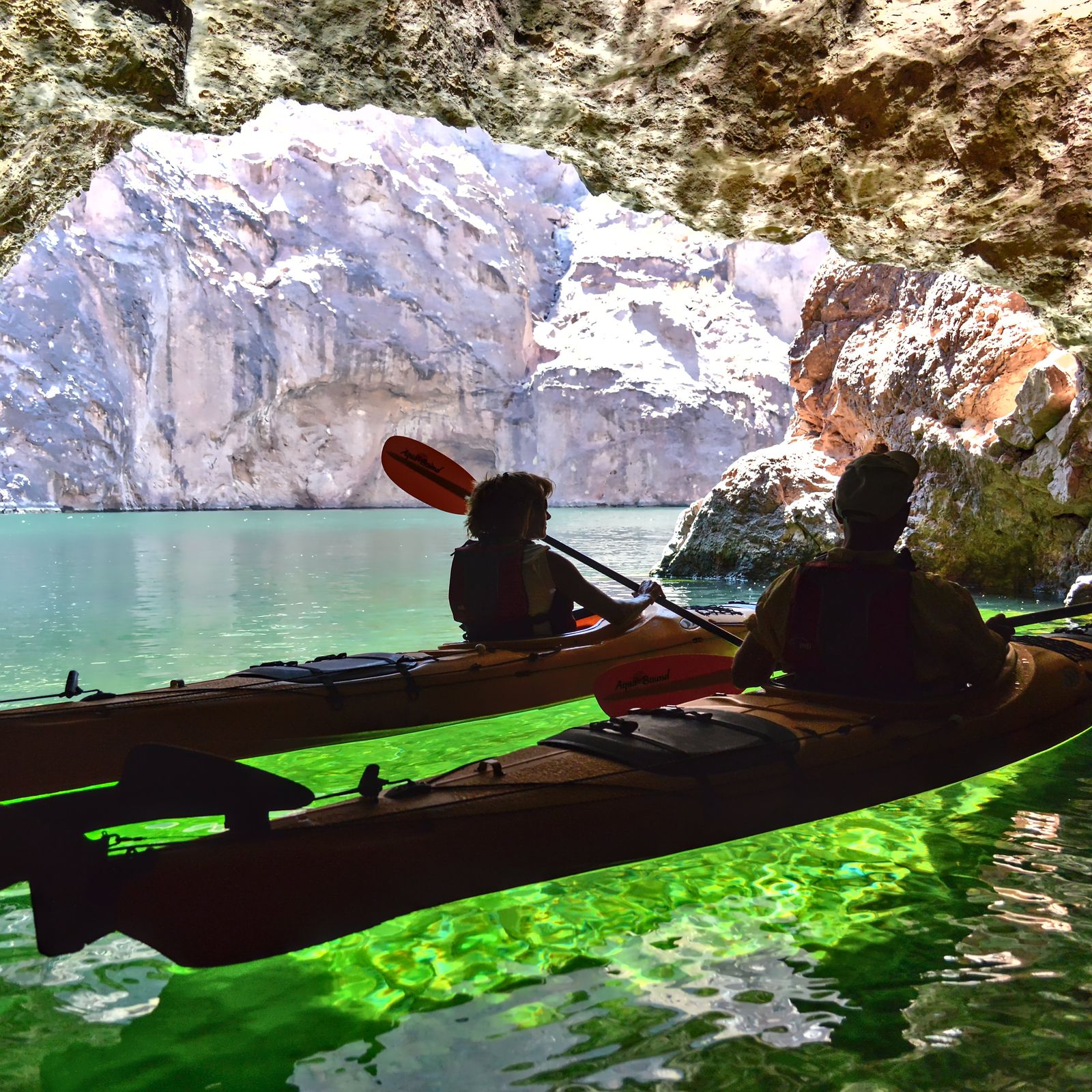 Two kayakers paddle inside a rocky cave with sunlight illuminating the green water.