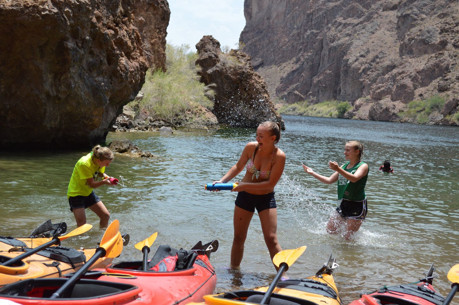 Three people playing with water near kayaks on a rocky riverbank.