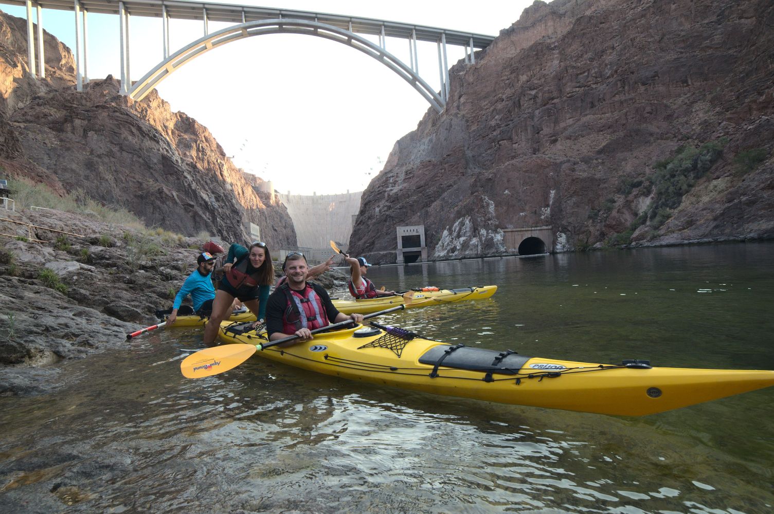 Group of kayakers in yellow kayaks on a river near a large bridge in a canyon.