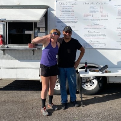 Two people smiling in front of a food truck, beside a small table with a bottle and plates.