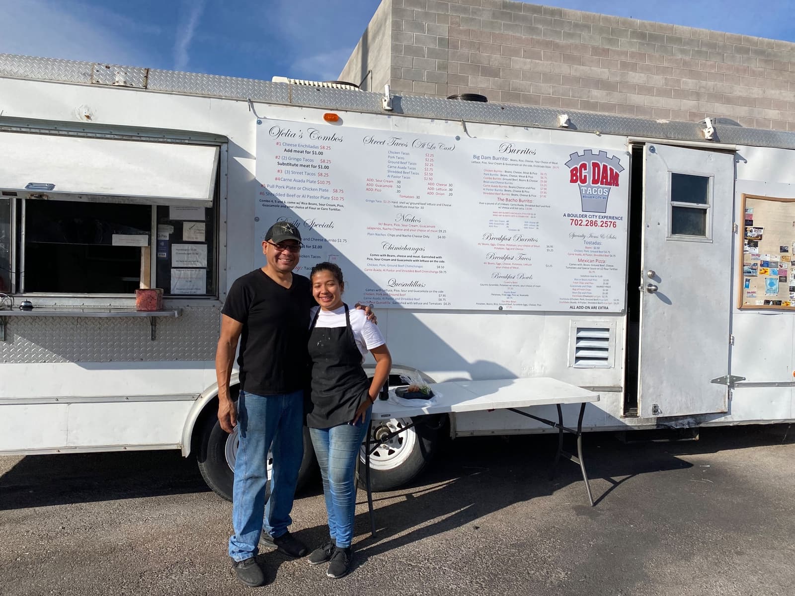 Two people stand together in front of a white food truck with menu and sign visible.