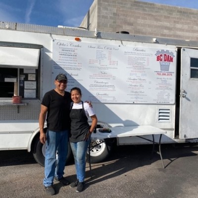 Two people smiling in front of a white food truck on a sunny day.