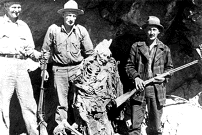 Three men with rifles stand next to a skeleton in tattered clothing against a rocky background.