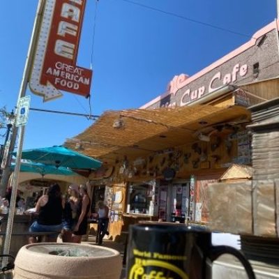 A coffee mug on a table outside a colorful cafe with a retro sign on a sunny day.