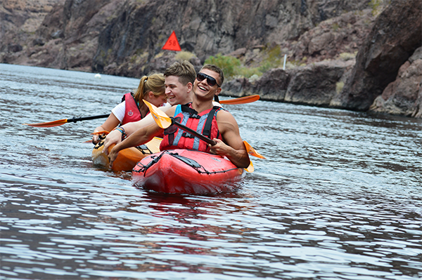 Three people kayaking on a river, wearing life jackets, with cliffs in the background.