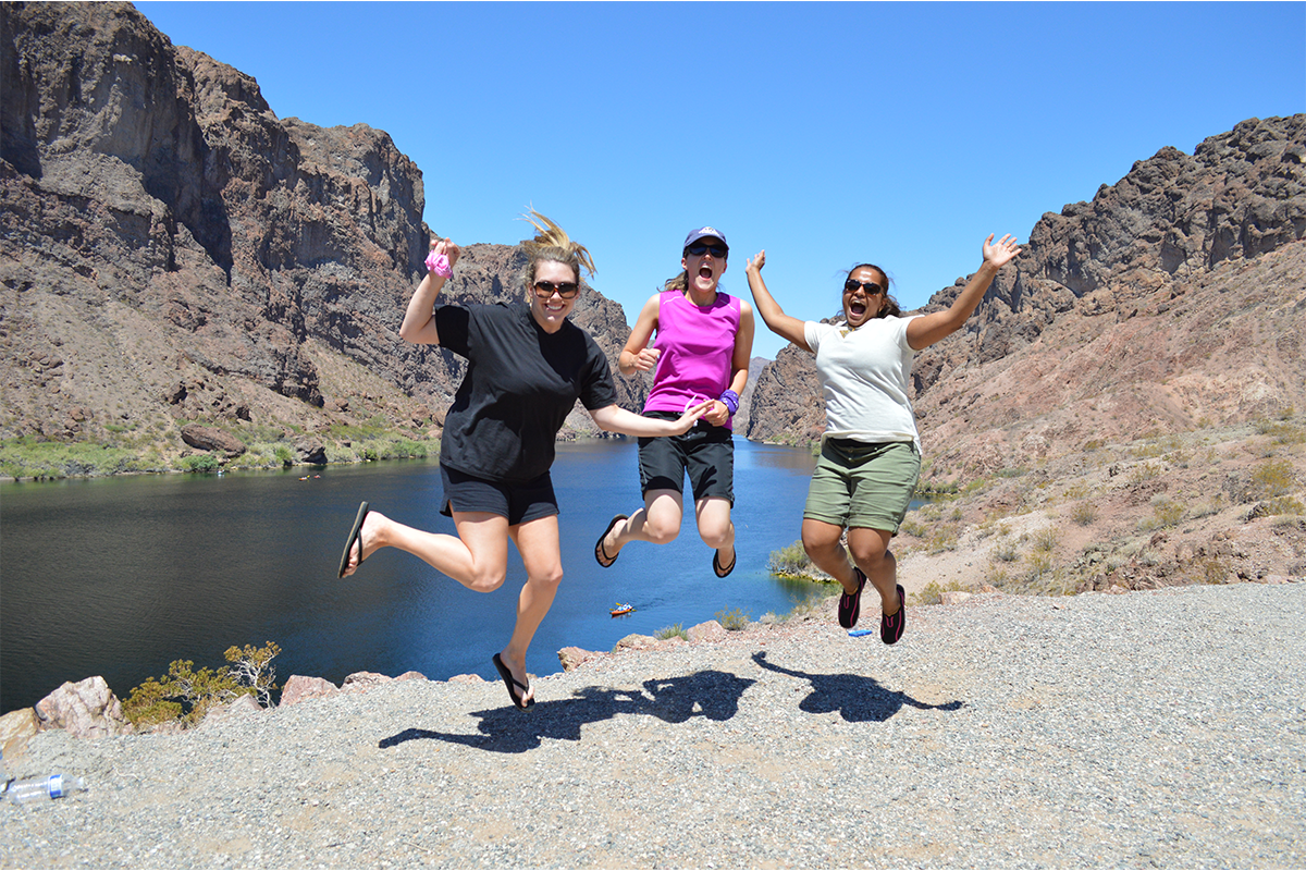 Three people jumping joyfully in front of a lake and rocky hills under a clear blue sky.