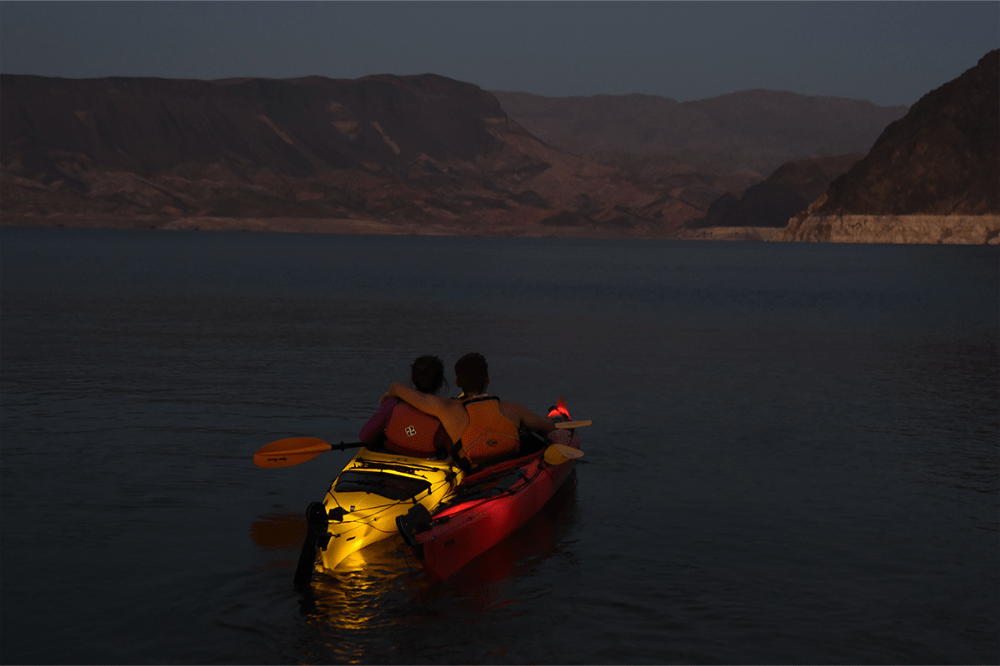 Two people in a kayak on a lake at dusk with mountains in the background, sitting close together.