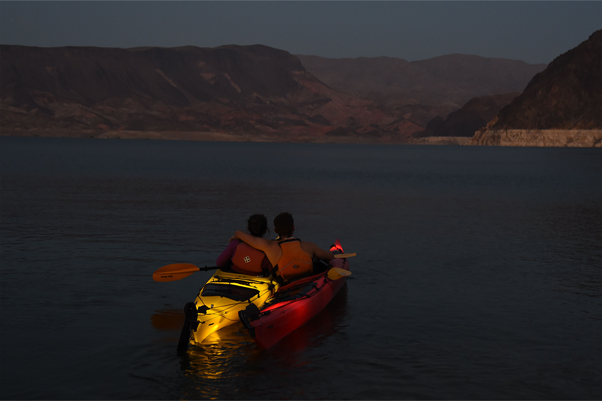 Two people in a kayak on a lake at dusk with mountains in the background, sitting close together.