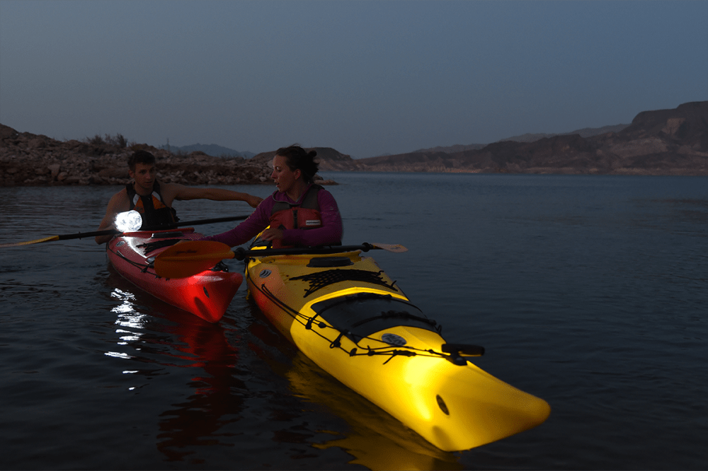 Two people kayaking at dusk with lights on their kayaks, near a rocky shoreline.