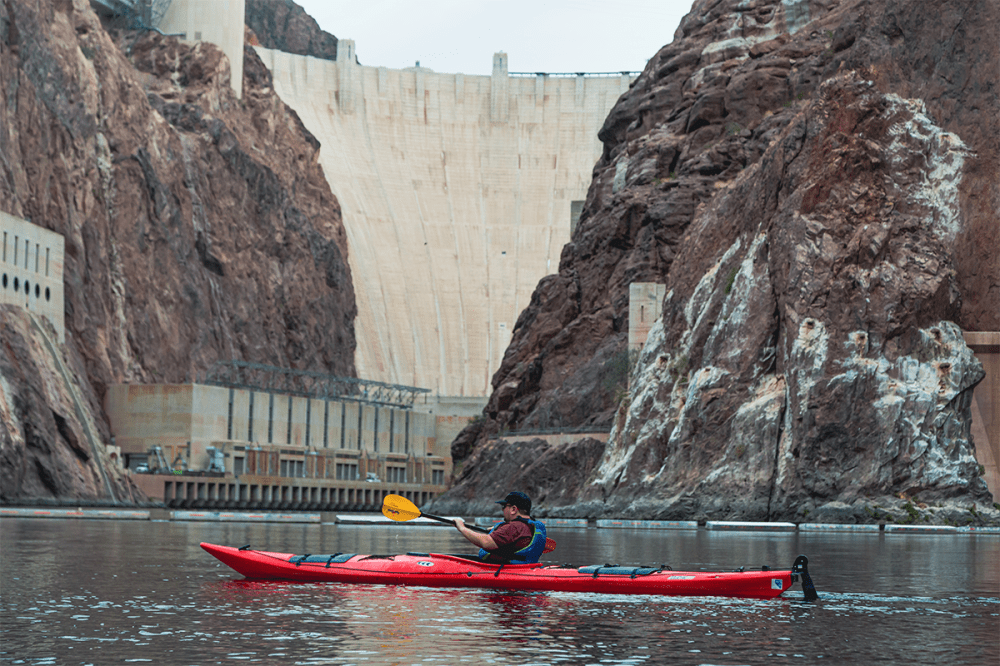 Person kayaking on a river with a large dam and rocky cliffs in the background.