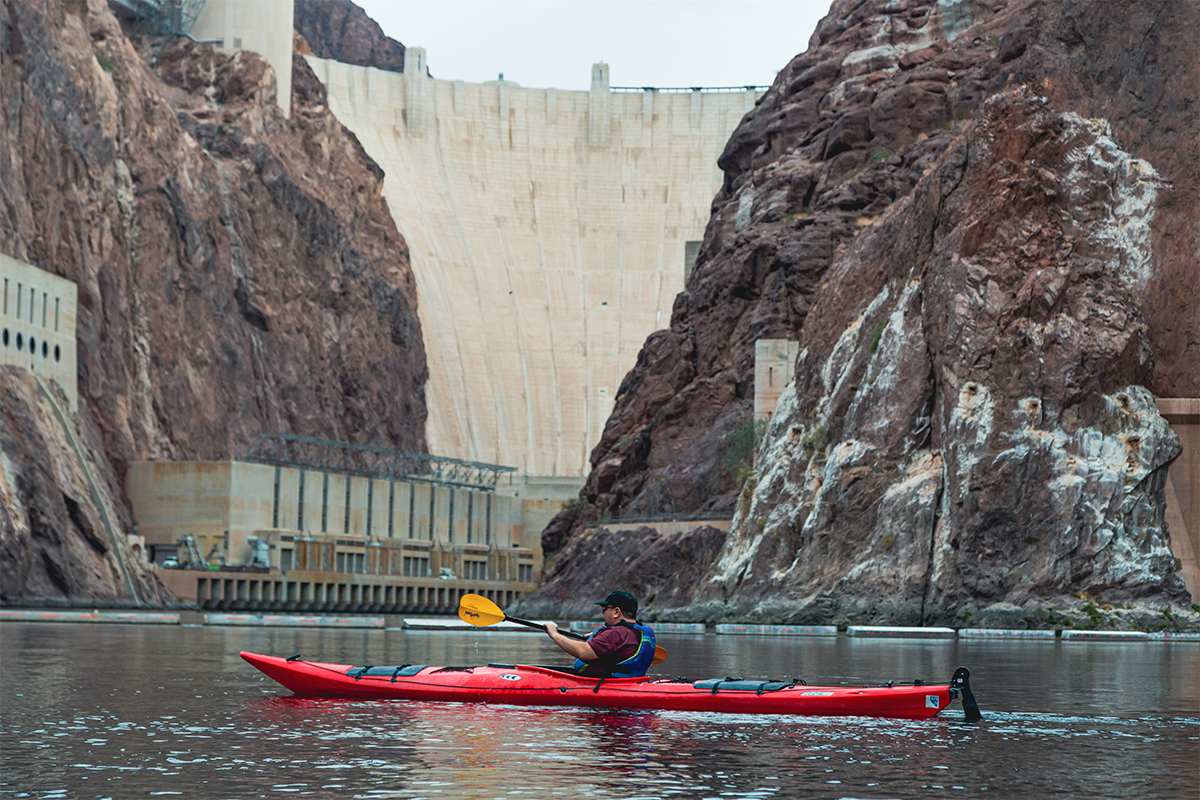 Person kayaking in front of a large dam and rocky cliffs.