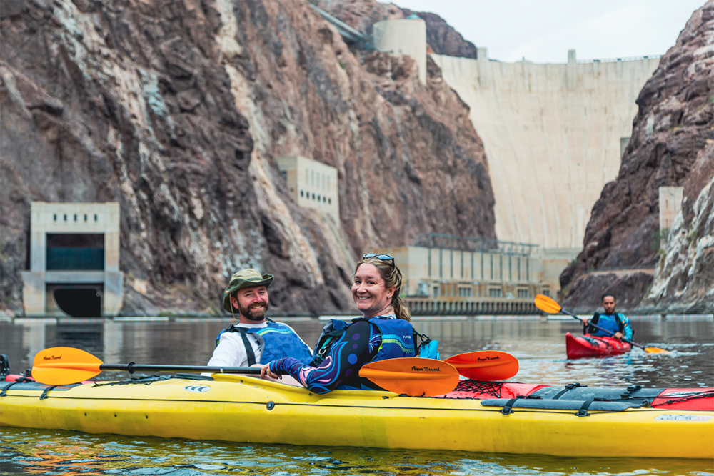 Two people kayaking on a river with a large dam in the background.