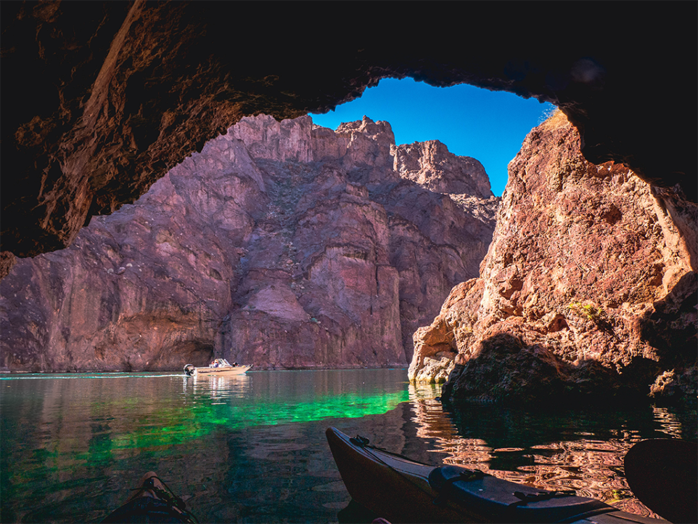 View from a cave of a boat on a clear lake surrounded by rocky cliffs under blue sky.
