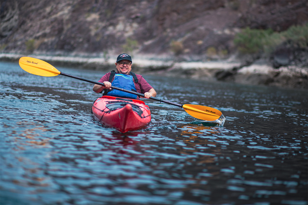 Person kayaking in a red kayak on a calm river, wearing a life jacket.